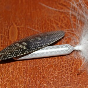 Op de afbeelding: Een zilveren Rooster Tail vislokmiddel met een witte body en een gevederde staart. Het lokmiddel heeft een textuur, schubachtig patroon op het metalen blad en de tekst "Rooster Tail" is zichtbaar. Ontworpen om vis te vangen.