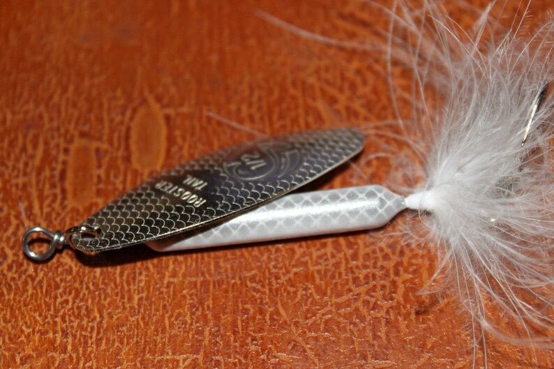 Op de afbeelding: Een zilveren Rooster Tail vislokmiddel met een witte body en een gevederde staart. Het lokmiddel heeft een textuur, schubachtig patroon op het metalen blad en de tekst "Rooster Tail" is zichtbaar. Ontworpen om vis te vangen.