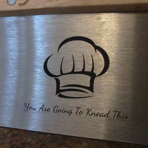 May include: A stainless steel pastry scraper with a chef's hat graphic and the phrase "You Are Going To Knead This" etched on the surface. The scraper has a brushed metal finish and is likely used for baking.
