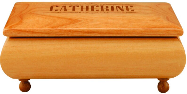 May include: A light-coloured wooden jewellery box with a rectangular shape and rounded edges. The lid is engraved with the name "CATHERINE". The box rests on four small, round feet.