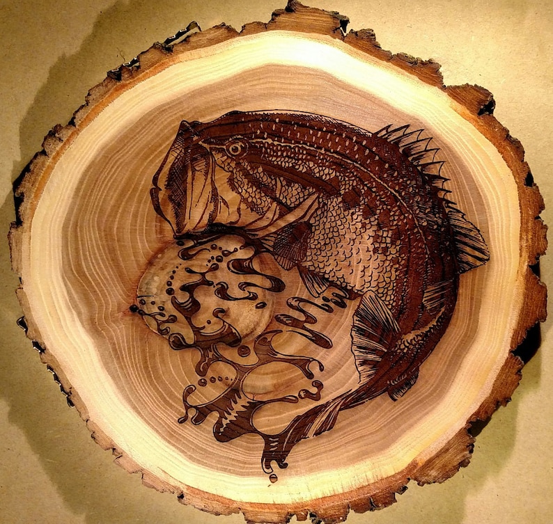 May include: A wood slice with a detailed pyrography design of a fish and water. The fish is brown with intricate scale and fin details. The wood has a natural light brown color with darker brown rings.