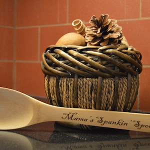 May include: A wooden spoon with the text "Mama's Spankin' Spoon" engraved on the handle. The spoon is next to a woven basket filled with decorative items, including a pine cone. The background features a brick wall.