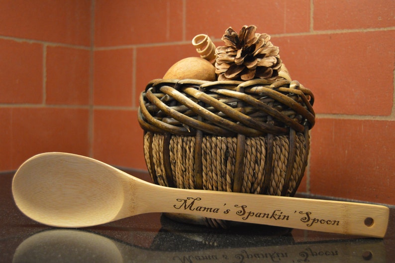 Op de afbeelding: Een houten lepel met de tekst "Mama's Spankin' Spoon" gegraveerd op de steel. De lepel ligt naast een gevlochten mand met decoratieve items, waaronder een dennenappel. De achtergrond is een bakstenen muur.