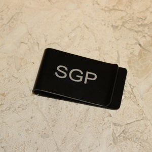 Pu&ograve; includere: Un fermasoldi in metallo nero con le iniziali "SGP" in lettere bianche. Il fermasoldi &egrave; rettangolare con un bordo curvo e poggia su una superficie chiara e decorata.
