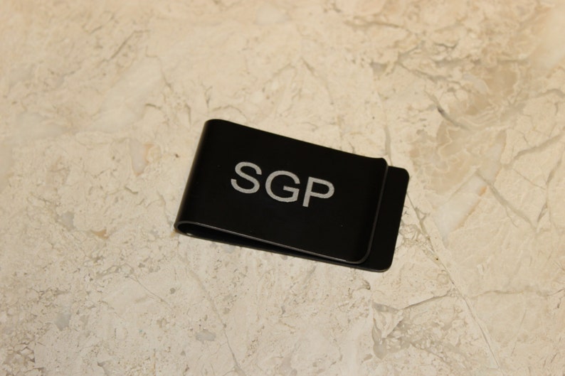 Pu&ograve; includere: Un fermasoldi in metallo nero con le iniziali "SGP" in lettere bianche. Il fermasoldi &egrave; rettangolare con un bordo curvo e poggia su una superficie chiara e decorata.