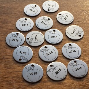 May include: A collection of silver-colored, circular metal tags with an apple design and the number "2015" etched on them. Each tag has a small hole at the top. The tags are scattered on a wooden surface.
