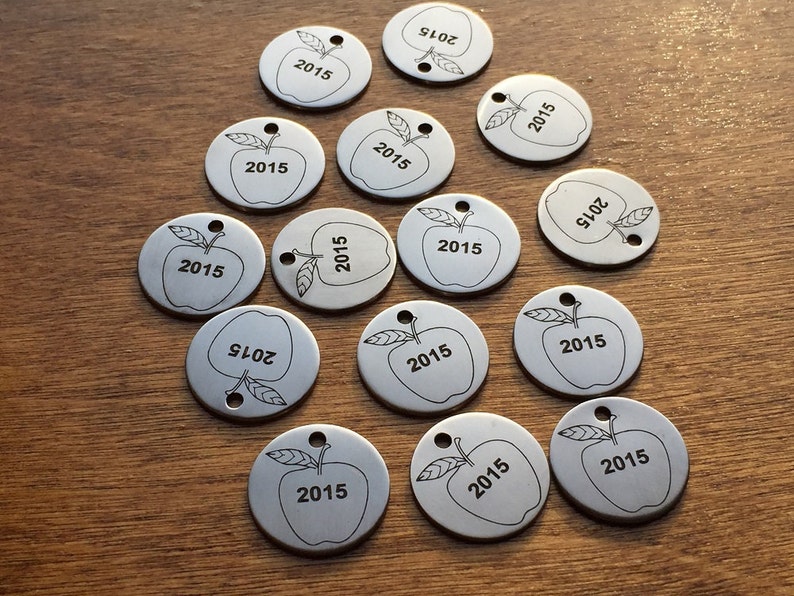 May include: A collection of silver-colored, circular metal tags with an apple design and the number "2015" etched on them. Each tag has a small hole at the top. The tags are scattered on a wooden surface.