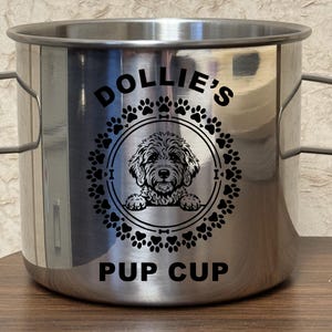 May include: A cylindrical stainless steel cup with two side handles. The front features a black graphic of a dog inside a circular frame of paw prints, with the text DOLLIE'S above the circle and the text PUP CUP printed below the circle.