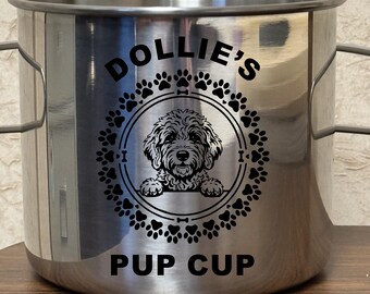 Op de afbeelding: Een cilindervormige roestvrijstalen beker met twee handvatten aan de zijkant. De voorzijde toont een zwarte afbeelding van een hond in een cirkelvormig kader van pootafdrukken, met de tekst DOLLIE'S erboven en PUP CUP eronder gedrukt.