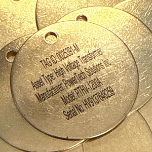 May include: Several circular metal tags with a hole at the top. The tags display the following text: TAG ID: 0025391-M, Asset Type: High Voltage Transformer, Manufacturer: PowerTech Solutions Inc., Model: PTHV-1200A, Serial No: HV9137648259.