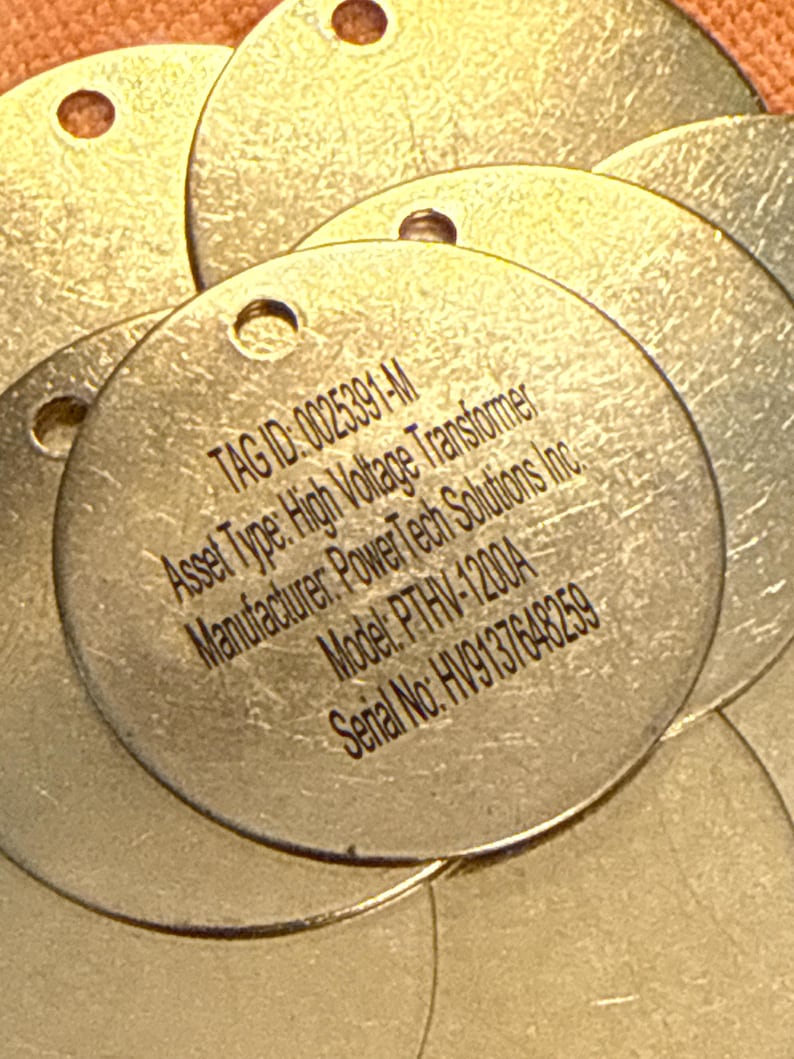 May include: Several circular metal tags with a hole at the top. The tags display the following text: TAG ID: 0025391-M, Asset Type: High Voltage Transformer, Manufacturer: PowerTech Solutions Inc., Model: PTHV-1200A, Serial No: HV9137648259.
