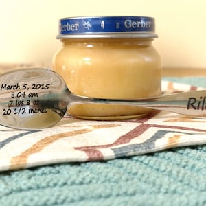 May include: A silver-toned metal spoon engraved with the name Riley on the handle and birth details on the bowl, including March 5, 2015, 8:04 am, 7 lbs 8 oz, and 20 1/2 inches, positioned in front of a small glass jar with a blue Gerber lid.