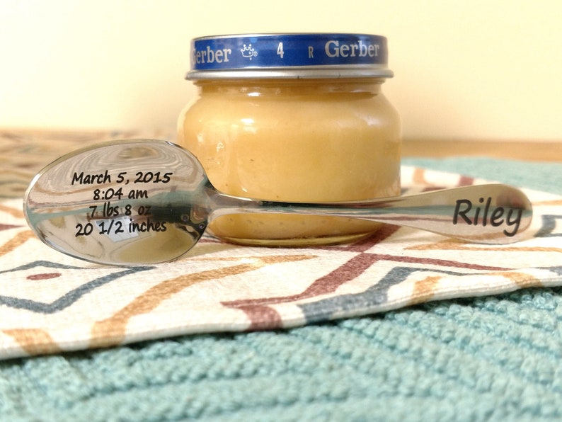 May include: A silver-toned metal spoon engraved with the name Riley on the handle and birth details on the bowl, including March 5, 2015, 8:04 am, 7 lbs 8 oz, and 20 1/2 inches, positioned in front of a small glass jar with a blue Gerber lid.