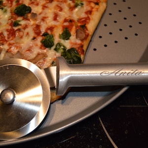 Op de afbeelding: Een roestvrijstalen pizzasnijder met de naam "Anita" gegraveerd op de handgreep, rustend op een geperforeerde pizzapan naast een pizzapunt met kaas en groene groenten.