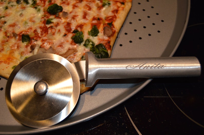 Op de afbeelding: Een roestvrijstalen pizzasnijder met de naam "Anita" gegraveerd op de handgreep, rustend op een geperforeerde pizzapan naast een pizzapunt met kaas en groene groenten.