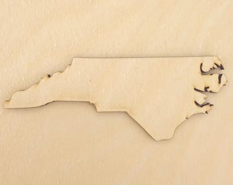 May include: A flat, laser-cut wooden cutout in the shape of the state of North Carolina. The piece features a light beige, natural wood grain finish with dark, burnt edges along the perimeter that define the state's geographic borders.