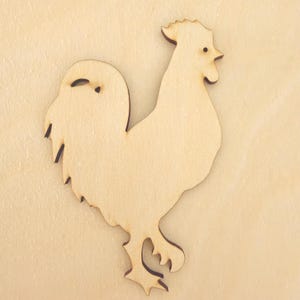 May include: A flat, light-coloured wooden cutout in the shape of a rooster. The silhouette features a detailed comb, a small circular eye, a pointed beak, and a large, textured tail. The surface shows natural wood grain patterns throughout the piece.