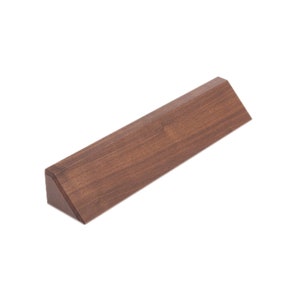 May include: A solid, triangular-shaped wooden object with a rich, dark brown finish. The piece has a flat top and a beveled edge, creating a geometric design. The wood grain is visible, adding texture to the minimalist design.