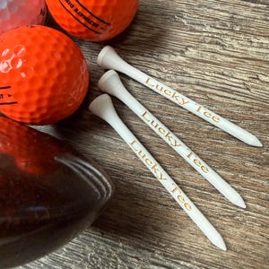 May include: Three white wooden golf tees with the text Lucky Tee printed in gold lettering, positioned next to two bright orange golf balls and a dark brown golf club head. The golf tees are arranged diagonally in a parallel formation.