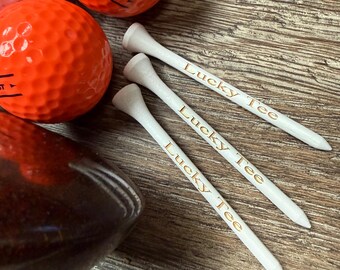 May include: Three white wooden golf tees with the text Lucky Tee printed in gold lettering, positioned next to two bright orange golf balls and a dark brown golf club head. The golf tees are arranged diagonally in a parallel formation.