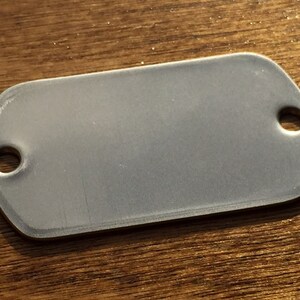 May include: A rectangular, silver metal tag with rounded corners and two holes. The tag has a matte finish. It is set against a wooden surface.
