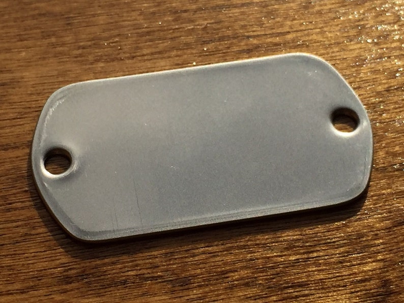 May include: A rectangular, silver metal tag with rounded corners and two holes. The tag has a matte finish. It is set against a wooden surface.