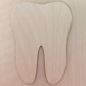 May include: A flat, light-colored wooden cutout in the shape of a human tooth. The object features a smooth, natural wood grain texture with a rounded top and two distinct roots at the bottom, presenting a simple, symmetrical silhouette.