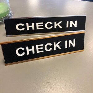May include: Two black rectangular signs with the words "CHECK IN" in bold white letters. One sign has a silver border, and the other has a gold border. The signs are on a light grey surface.