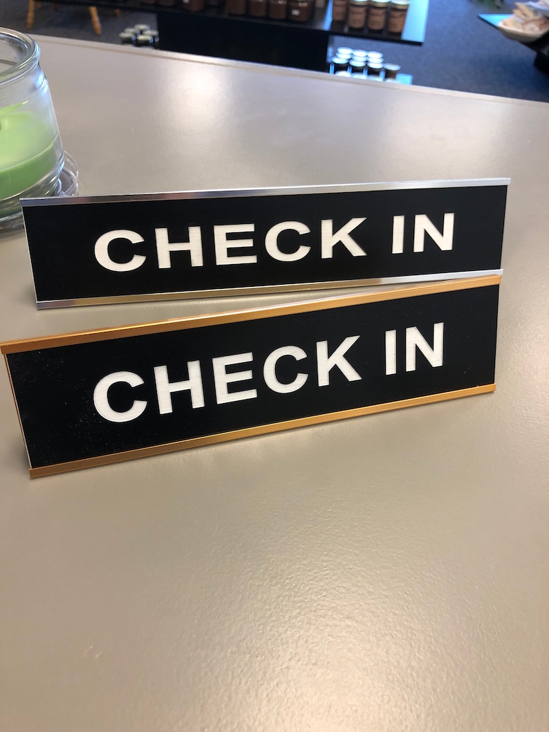 May include: Two black rectangular signs with the words "CHECK IN" in bold white letters. One sign has a silver border, and the other has a gold border. The signs are on a light grey surface.
