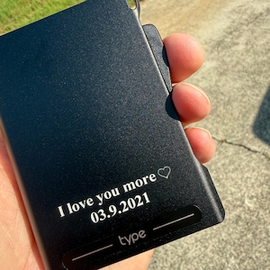 Może przedstawiać: Prostokątny wizytownik z matowego czarnego metalu z wygrawerowanym laserowo napisem I love you more, symbolem serca oraz datą 03.9.2021. W dolnej części znajduje się słowo type umieszczone pomiędzy dwiema poziomymi liniami.