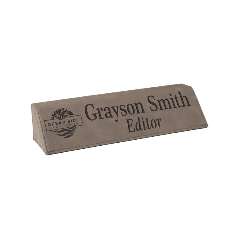 May include: A gray desk nameplate with the text "Grayson Smith Editor" in a serif font. The nameplate also features the logo of "Ocean Side Publishing Company". The nameplate is made of a textured material and has a triangular shape.