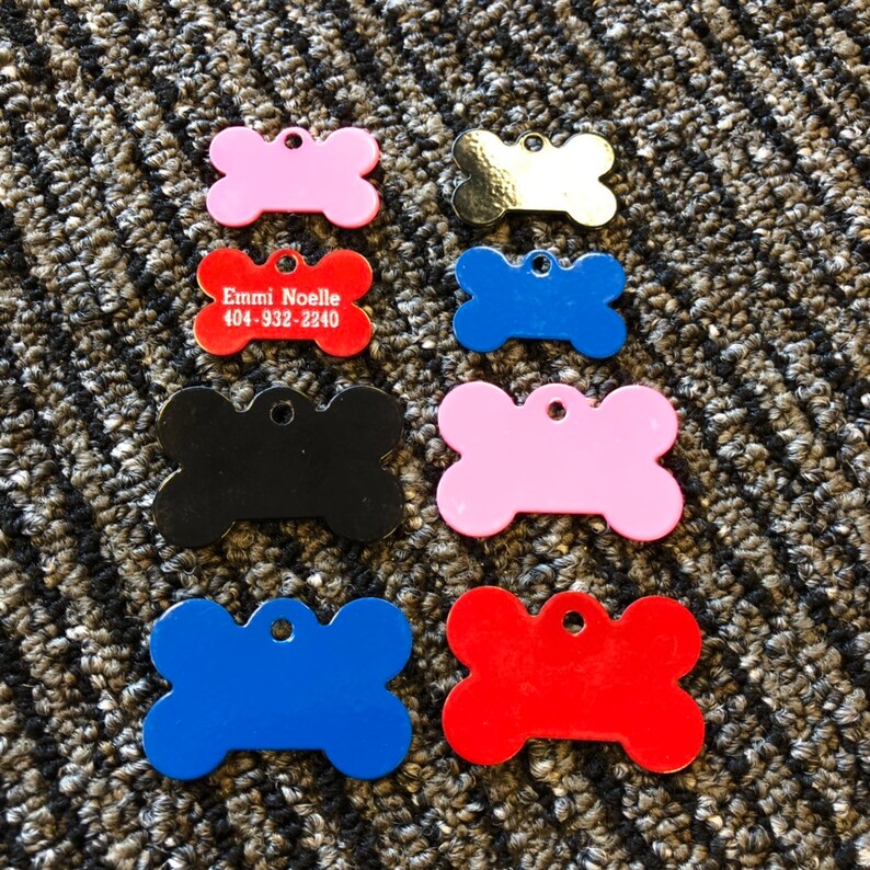 Puede incluir: Una colecci&oacute;n de etiquetas de identificaci&oacute;n para mascotas en forma de hueso en varios colores, incluyendo rosa, rojo, azul, negro y dorado. Una etiqueta roja muestra el nombre "Emmi Noelle" y un n&uacute;mero de tel&eacute;fono. Cada etiqueta tiene un agujero para sujetar.