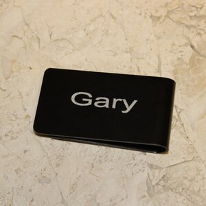 Pu&ograve; includere: Un fermasoldi nero con il nome "Gary" in lettere bianche. La clip &egrave; rettangolare con bordi arrotondati e un design curvo. Lo sfondo &egrave; una superficie chiara e decorata.