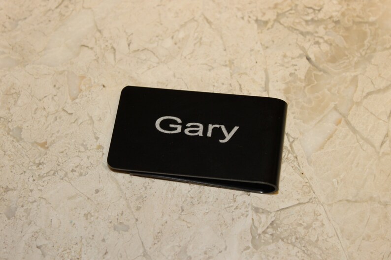 Pu&ograve; includere: Un fermasoldi nero con il nome "Gary" in lettere bianche. La clip &egrave; rettangolare con bordi arrotondati e un design curvo. Lo sfondo &egrave; una superficie chiara e decorata.