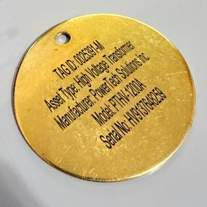 May include: A circular, gold-toned metal tag with a small hole at the top. The tag features black text: "TAG ID: 0025391-M, Asset Type: High Voltage Transformer, Manufacturer: PowerTech Solutions Inc., Model: PTHV-1200A, Serial No: HV9137648259."
