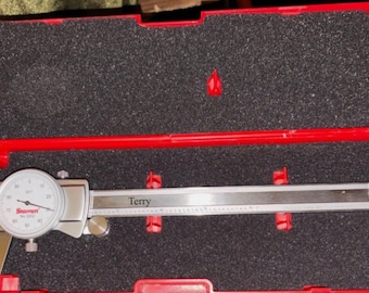 May include: A silver metal dial calliper with a white face and black markings, featuring the name Terry printed on the beam. The tool is housed inside a bright red plastic storage case with black foam padding and red plastic support clips.