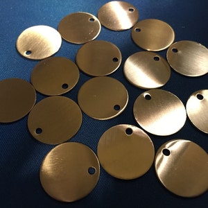 May include: A collection of small, round, brass-colored metal tags, each with a small hole near the edge. The tags have a brushed finish and are scattered across a blue surface. These tags are suitable for jewelry making or crafting.