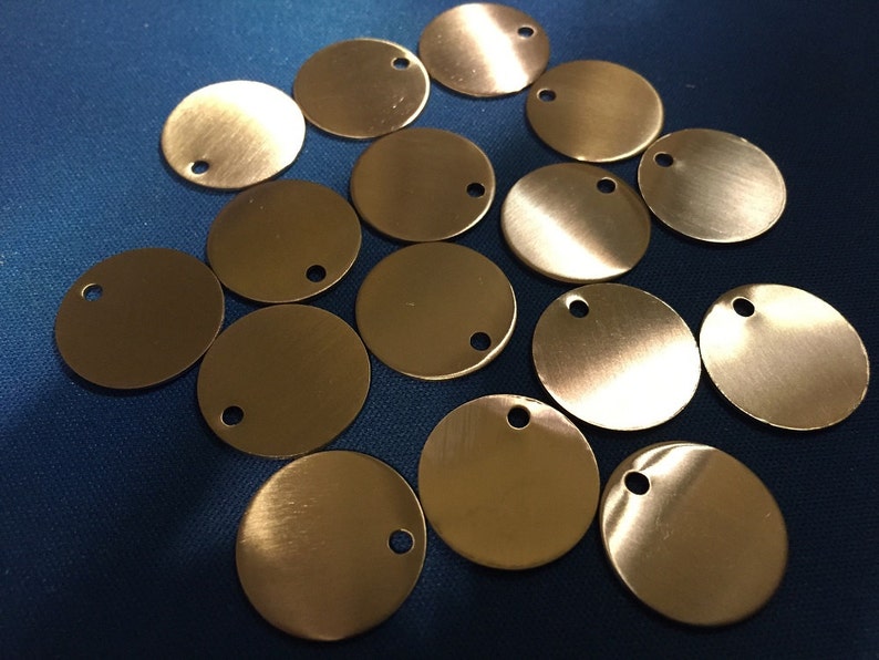 May include: A collection of small, round, brass-colored metal tags, each with a small hole near the edge. The tags have a brushed finish and are scattered across a blue surface. These tags are suitable for jewelry making or crafting.