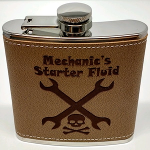 May include: A stainless steel flask wrapped in tan textured material with white stitching. The front features an engraved design of two crossed wrenches above a skull and crossbones, with the text "Mechanic's Starter Fluid" engraved above.