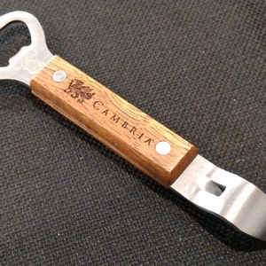 May include: A bottle opener with a stainless steel head and a wooden handle. The handle is engraved with the word "CAMBRIA" and a decorative emblem. The opener is designed for both bottle caps and can lids.