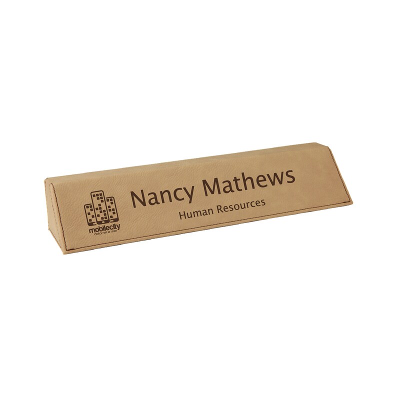 May include: A light brown desk nameplate with the name "Nancy Mathews" and the title "Human Resources" engraved on it. The nameplate also features the "mobilecity" logo. The nameplate is a triangular prism shape.
