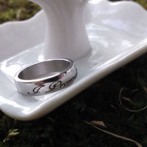 May include: A silver ring with the inscription "I Promise" rests on a white, scalloped-edge dish. The ring is set against a backdrop of green moss, creating a natural and elegant presentation. The ring is a symbol of commitment.
