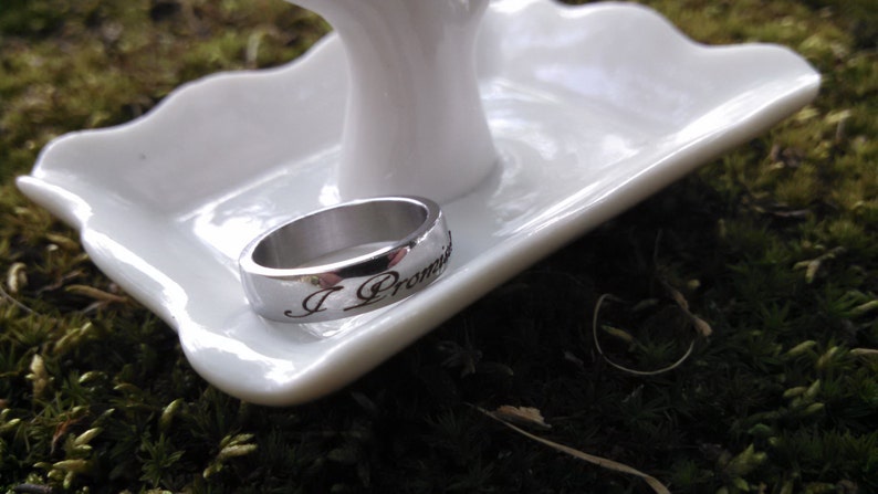 May include: A silver ring with the inscription "I Promise" rests on a white, scalloped-edge dish. The ring is set against a backdrop of green moss, creating a natural and elegant presentation. The ring is a symbol of commitment.