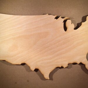 Puede incluir: Un recorte de madera de color claro con la forma de los Estados Unidos contiguos. La superficie muestra un patrón de veta de madera natural con bordes lisos y lijados que siguen el contorno geográfico del país de costa a costa.