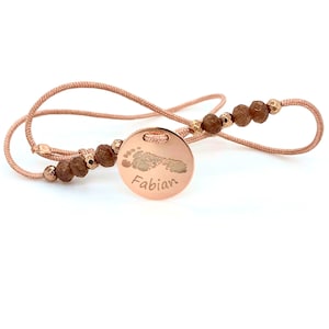 Könnte beinhalten: Halskette aus Roségold mit einem runden Anhänger mit einem Babyfußabdruck und dem Namen "Fabian". Die Halskette hat eine feine Kordel mit braunen und goldenen Perlen.