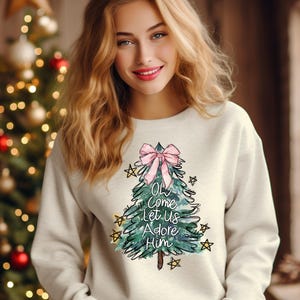 May include: White sweatshirt with a green watercolor Christmas tree design. The tree has a pink bow and gold stars. The text on the tree reads "Oh Come Let Us Adore Him".