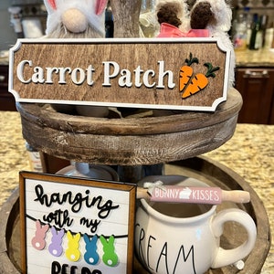 May include: A tiered wooden tray with a wooden sign that says "Carrot Patch" and a wooden sign that says "hanging with my peeps" with a string of colorful peeps hanging from it. There is a white mug with the word "CREAM" on it and a pink rolling pin with the words "Bunny Kisses" on it.