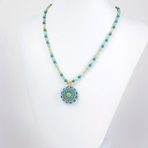 May include: A turquoise and gold beaded necklace with a turquoise and pink beaded pendant. The pendant is a circular design with a turquoise stone in the center.