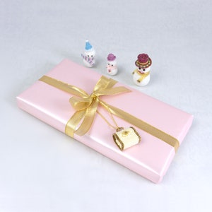 May include: A pink gift box with a gold ribbon and bow. A small white beaded purse hangs from the ribbon. Three small snowman ornaments are on the surface next to the box.