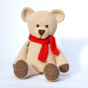 May include: A handmade, beige crochet teddy bear with a red scarf. The bear has brown eyes, a brown nose, and brown paws. The bear is sitting upright against a white background.